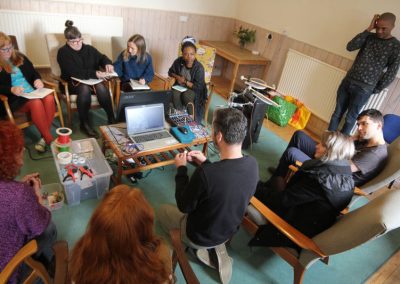 A group of individuals sitting around a table, working together to build something reusing bits of technology. The table holds a laptop and a sound controller.