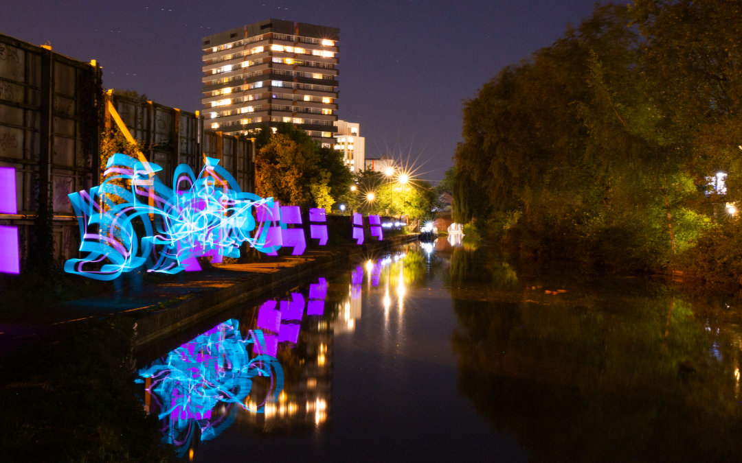 The Coventry Canal at night. A block of flats and some streetlamps glow in the middle-distance, but the water and the towpath are dark. Some trees hang over the canal on the right side while on the towpath to the left, ribbons of blue light flow along its length, punctuated by purple semi-colons: The Random String Festival logo.