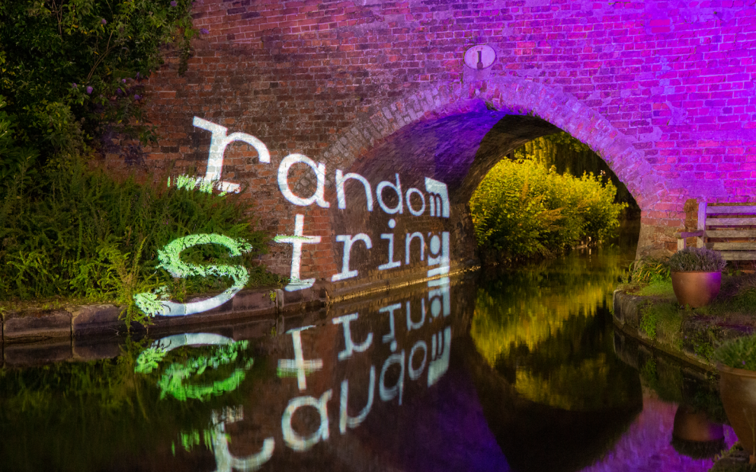 Bridge 1 at the end of Coventry Canal Basin, nighttime. The canal flows under the old brick bridge and into the dark distance. The bridge itself is lit with a bright pink glow, the words 'random string' projected onto it in white, lighting up the texture of the brickwork and reflecting in the water below.