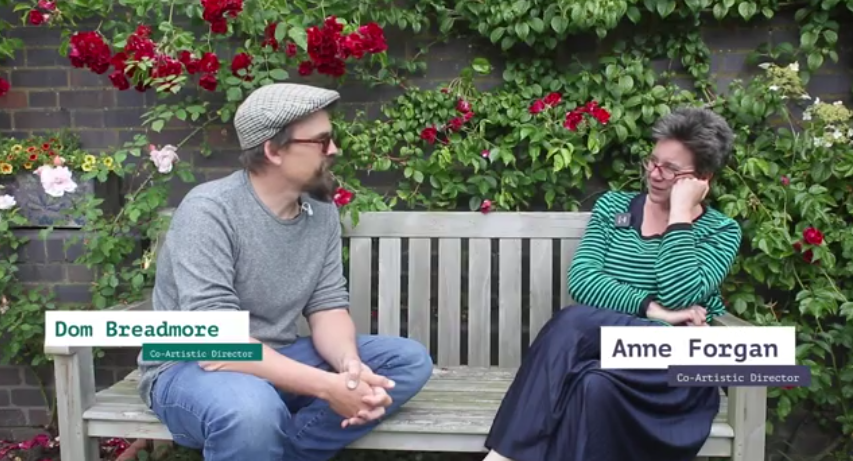 Co-Artistic Directors, Dom Breadmore & Anne Forgan, sit on a wooden bench in a walled garden with red roses. Dom is a white man in his late thirties, he had a dark moustache and goatie and wears a grey jumper and jeans with a grey flatcap and glasses. Anne is a white woman in her fifties, she has short, dark hair and wears a striped green jumper and navy skirt, she also wears glasses.